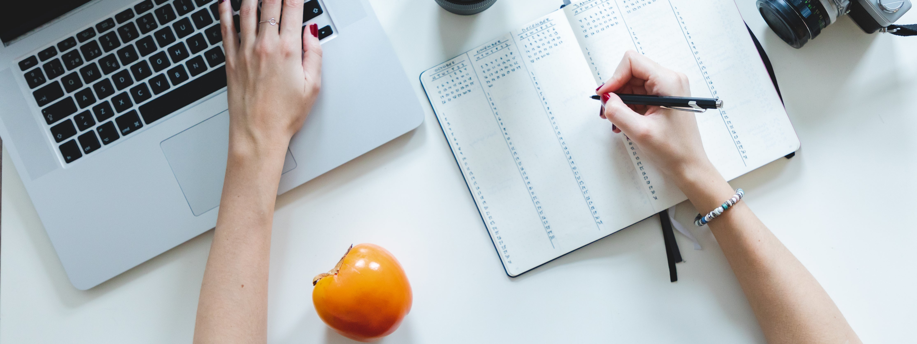 Person using a laptop with a planner and an orange fruit on a desk